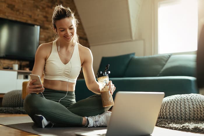 Young happy athletic woman relaxing on the floor and using laptop while drinking smoothie at home.