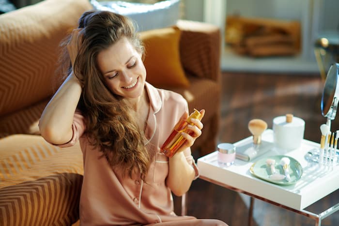happy stylish 40 years old woman in pajamas with long brunette wavy hair holding hair oil bottle near table with toiletries in the modern living room in sunny winter day.