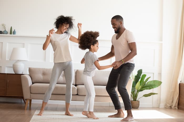 Happy funny active african family with cute little kid daughter dancing at home, carefree cheerful black parents mom dad and small child girl having fun jumping laughing enjoy leisure in the morning