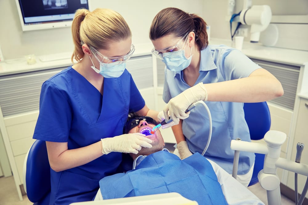 people, medicine, stomatology and health care concept - female dentist and assistant with dental curing light and mirror treating male patient teeth at dental clinic office