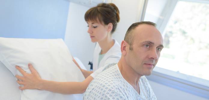 A male patient sitting up in his bed while a nurse fluffs up a pillow behind him in an attempt to help him feel more comfortable