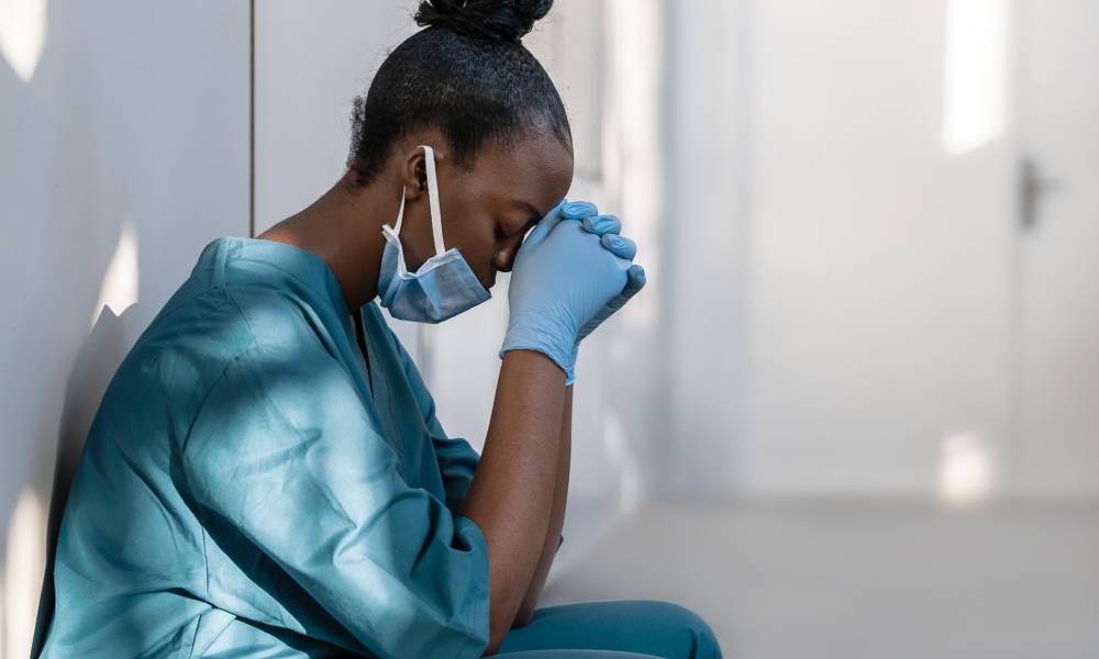 An exhausted and depressed female healthcare worker crouches against a hospital wall with her head down.