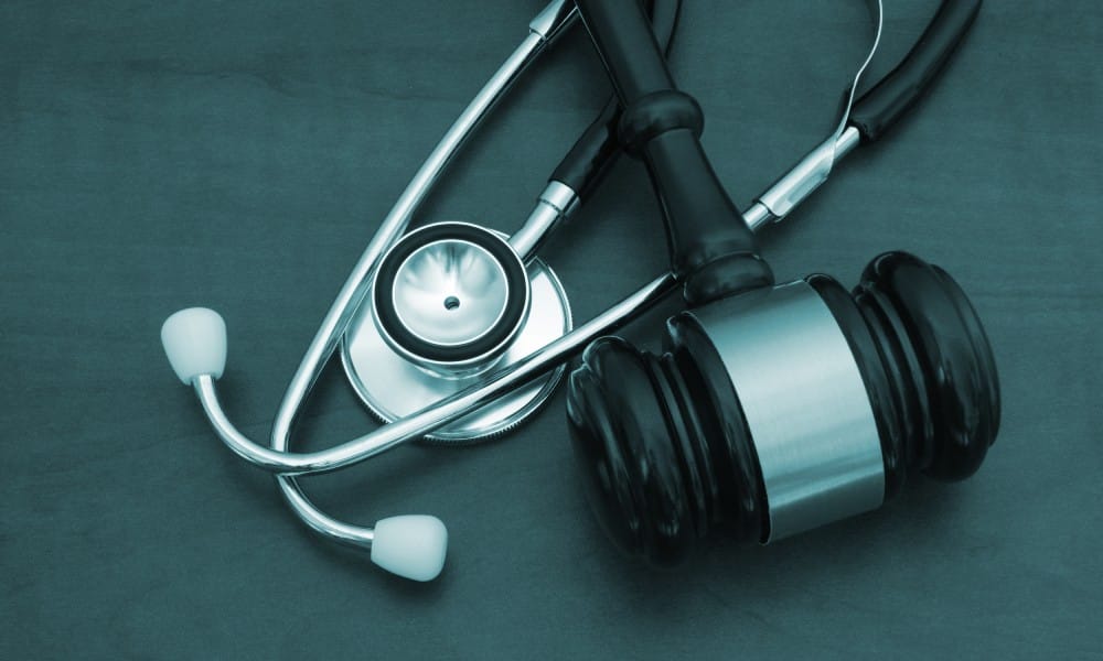 A stethoscope with a black gavel laid on top of it on a dark teal wooden background.
