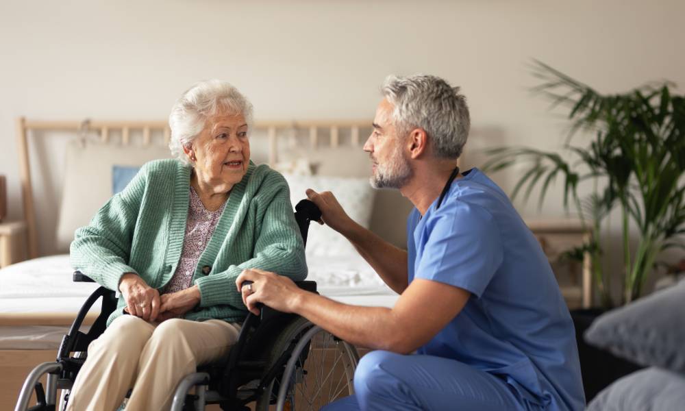 A male medical professional in light blue scrubs visiting with his older female patient as she sits in a wheelchair at home.
