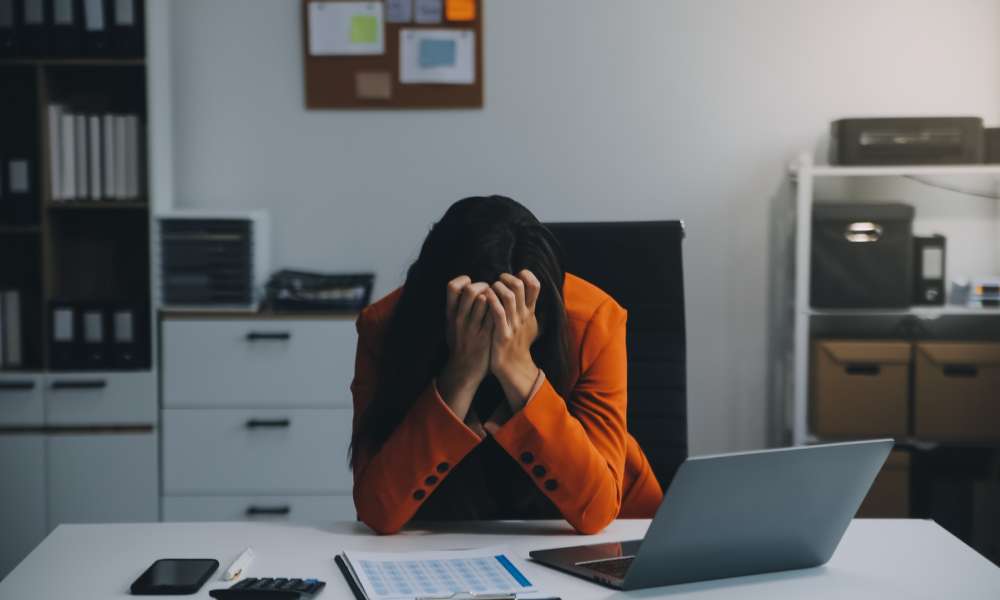 A stressed-out female employee has her head resting in her hands at her desk. She's wearing an orange blazer.