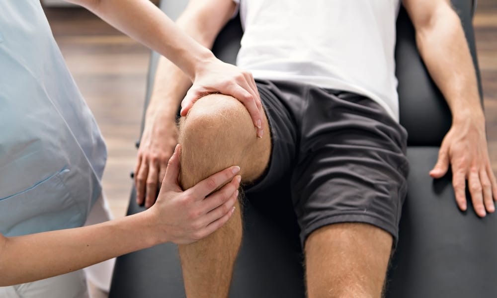 A man in gray shorts is undergoing a physical therapy session. His therapist is aiding him in lifting his knee up.