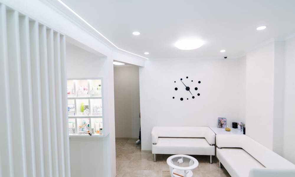 An empty waiting room contains two white couches, a small white coffee table, wooden floors, and a black clock.