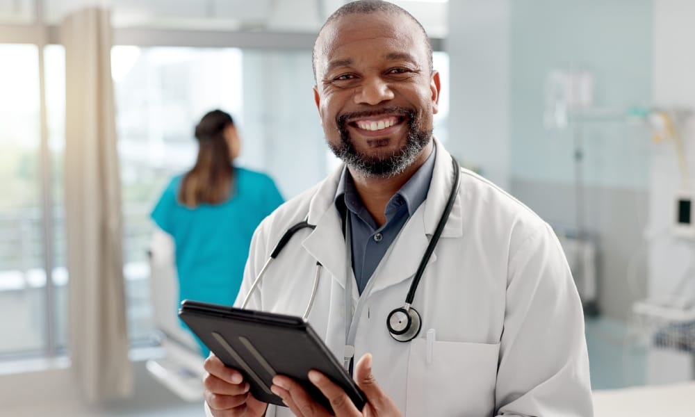 A middle-aged male doctor smiling widely and warmly at the camera. He is wearing a white coat and holding a tablet.