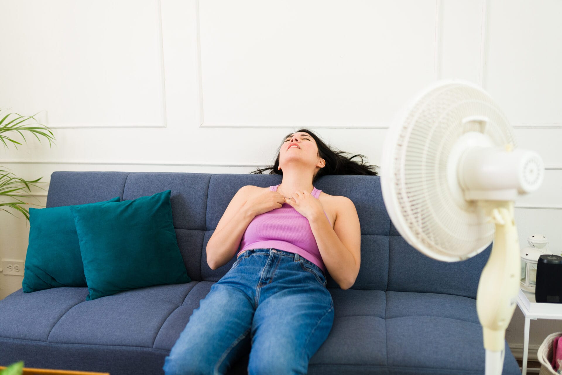 An uncomfortable woman sitting on her couch with a rotating standing fan in front of her. She is trying to cool off.