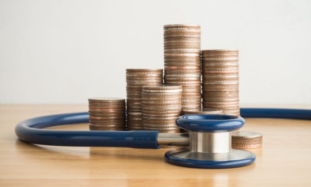 A stethoscope lying are a table, surrounding a large pile of change. The change is piled nicely into stacks of coins.