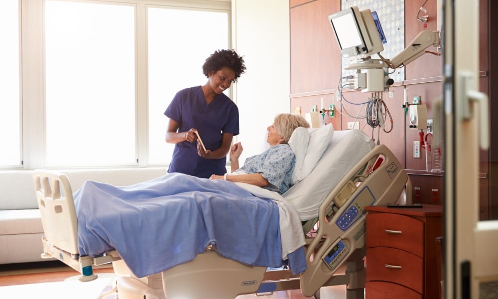 A young woman in navy-blue scrubs holds a tablet as she talks to a grey-haired patient lying in a hospital bed.