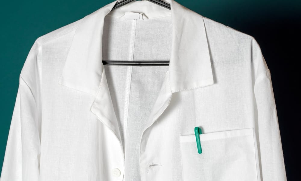 A lab coat on a hanger that is unbuttoned. The hanger is black, and there is a green pen in the pocket square.