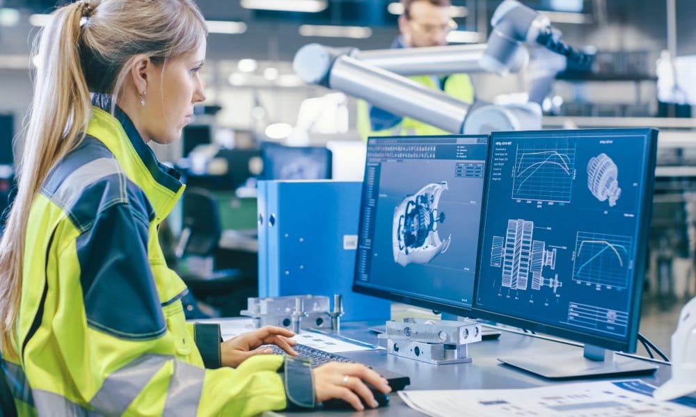 Young female engineer wearing a safety jacket, using a desktop computer displaying a 3D model of a product inside a warehouse.