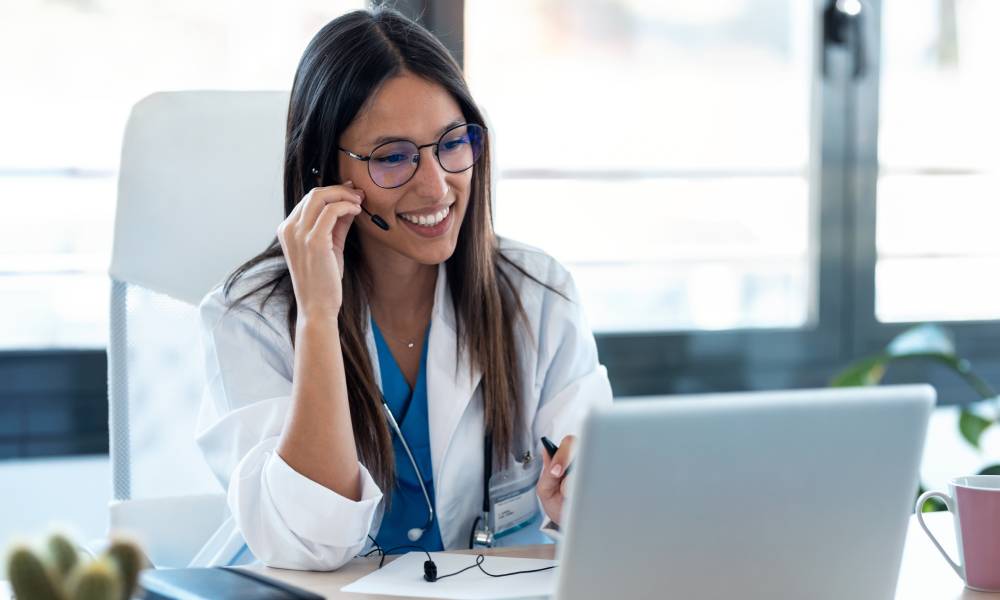 A female healthcare provider sits at a desk, smiling at an open laptop. She adjusts the microphone on her headset.