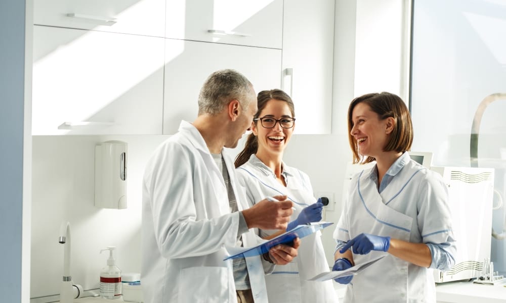 Three medical professionals wearing medical scrubs and some gloves laugh with each other in a medical office.