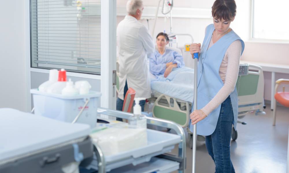A custodian in a blue smock sweeping the floor just outside a patient's room. Behind her, the patient and doctor are talking.