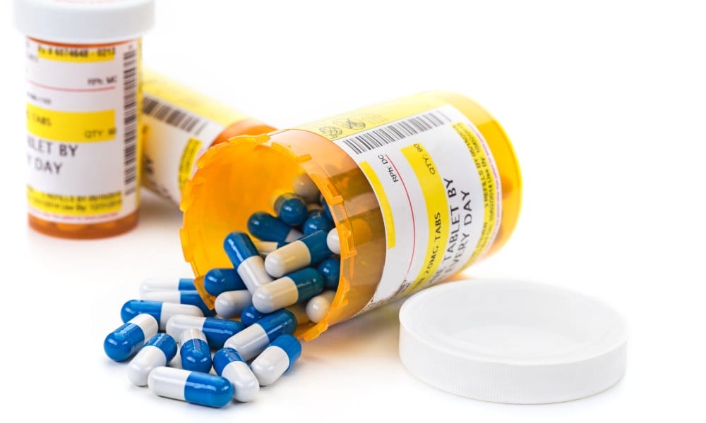 Three orange pill bottles sit next to each other. One of them has tipped over and has blue and white pills spilling out.