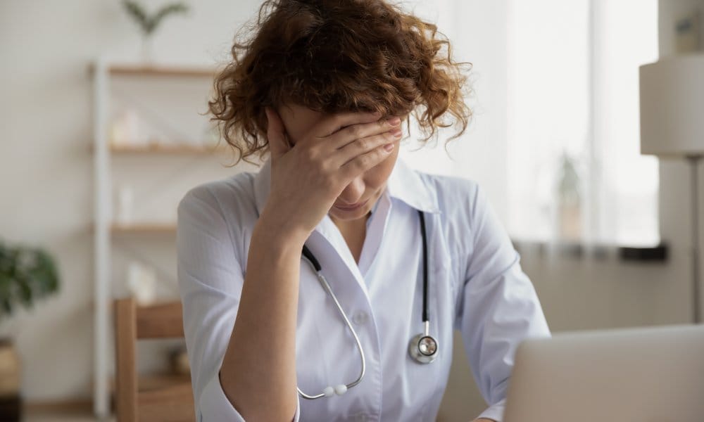 A female physician visibly frustrated with her head in her hand. She's sitting down at a table with a laptop in front of her.