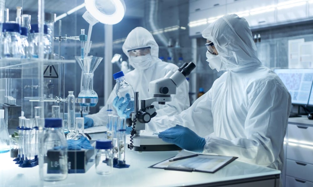Two people in full-coverage PPE masks, glasses, suits, and gloves conducting research in a bright lab with tubes and beakers.