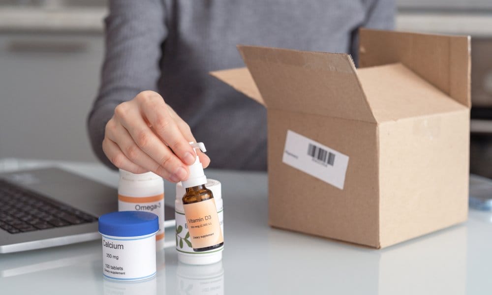 A woman at a table is taking various health supplements in bottles and containers out of a cardboard shipping box.