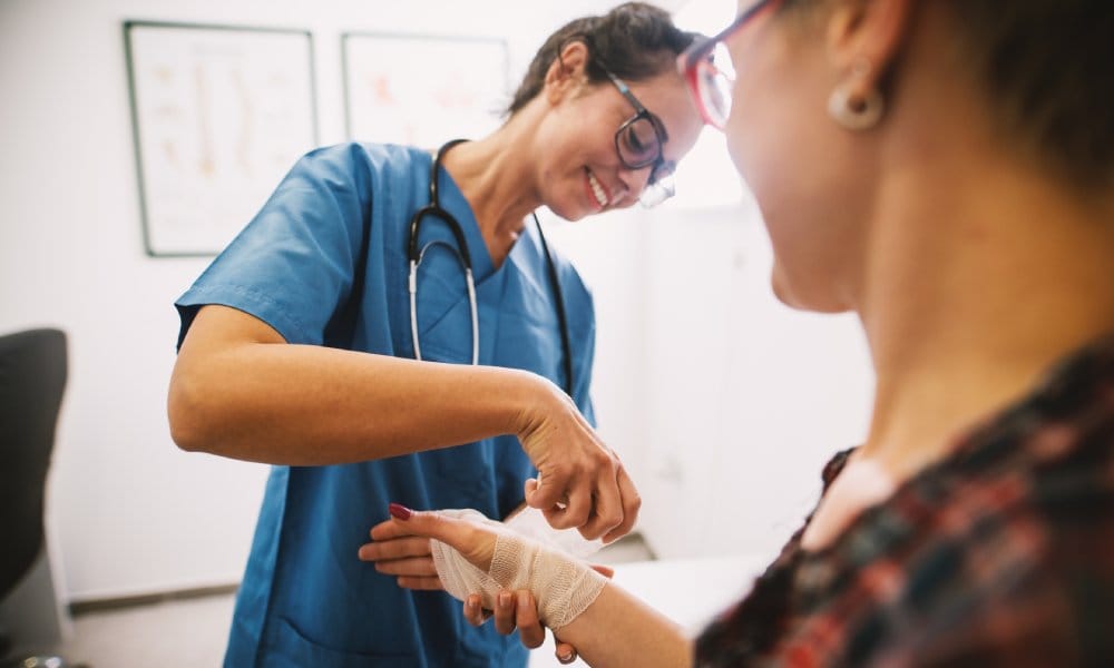 A female medical professional in teal scrubs applies a bandage to a patient's hand. The patient is sitting.