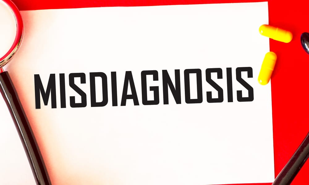 A red background featuring a paper with "misdiagnosis" written in bold letters, with a stethoscope and pills resting on top.