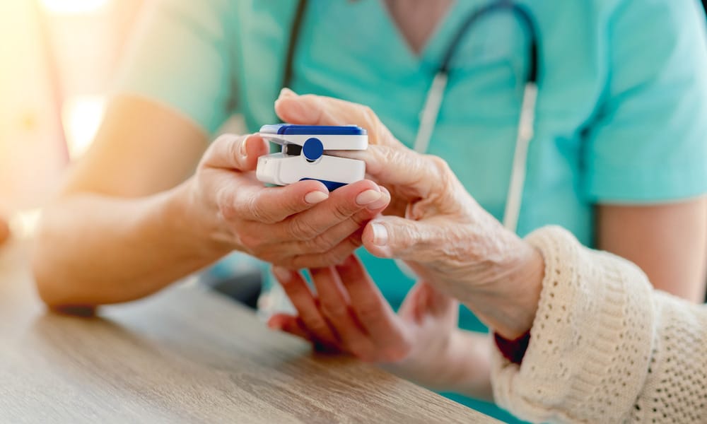 A healthcare professional in teal scrubs places a pulse oximeter on the hand of an older patient in a tan sweater.