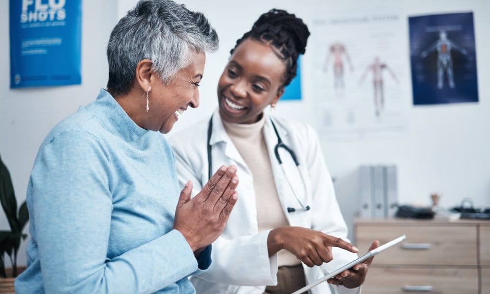 A woman smiling and pressing her palms together in gratitude as a doctor points to something on a tablet in front of them.