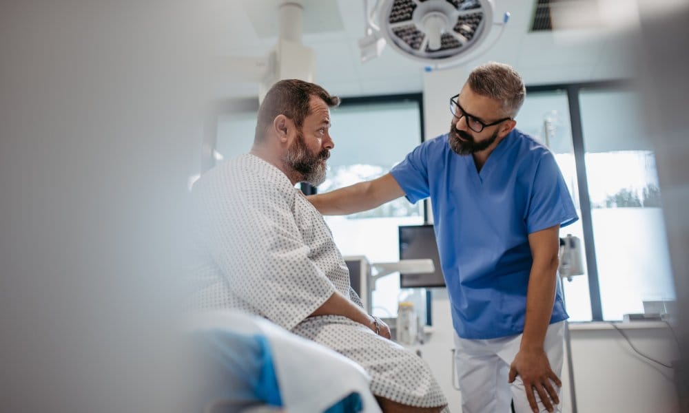 A doctor in dark blue scrubs placing a hand on a bearded patient’s shoulder as a gesture of comfort.