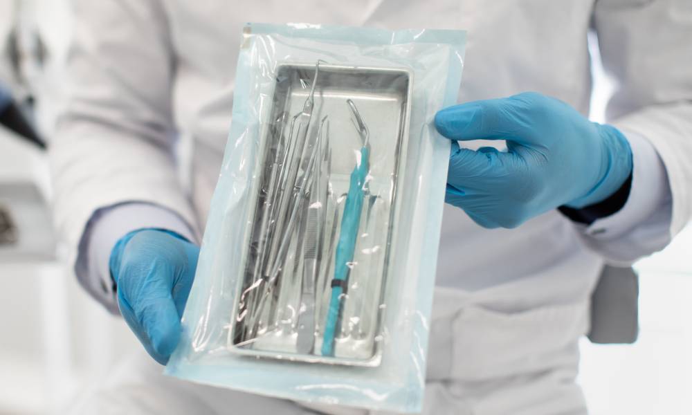 A healthcare professional wearing blue gloves and holding a plastic sterilization pouch of metal dental instruments.
