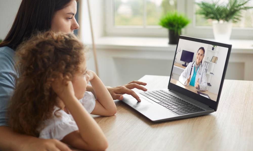 A mother and her child sitting at the dining room table on a telehealth video call with a primary care provider.