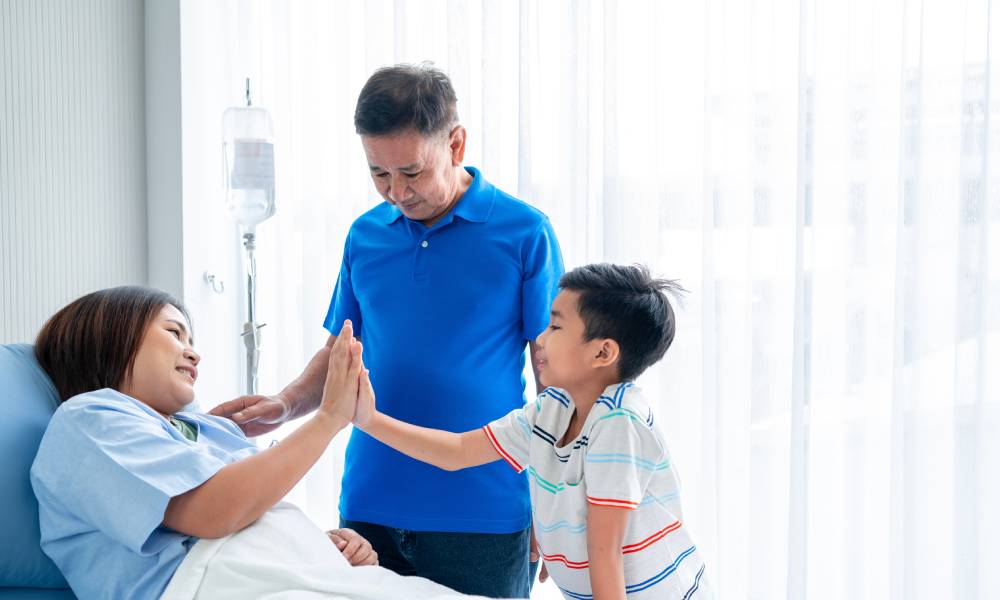 A woman in a hospital gown laying on a hospital bed while a young boy gives her a gentle high five and a man watches.
