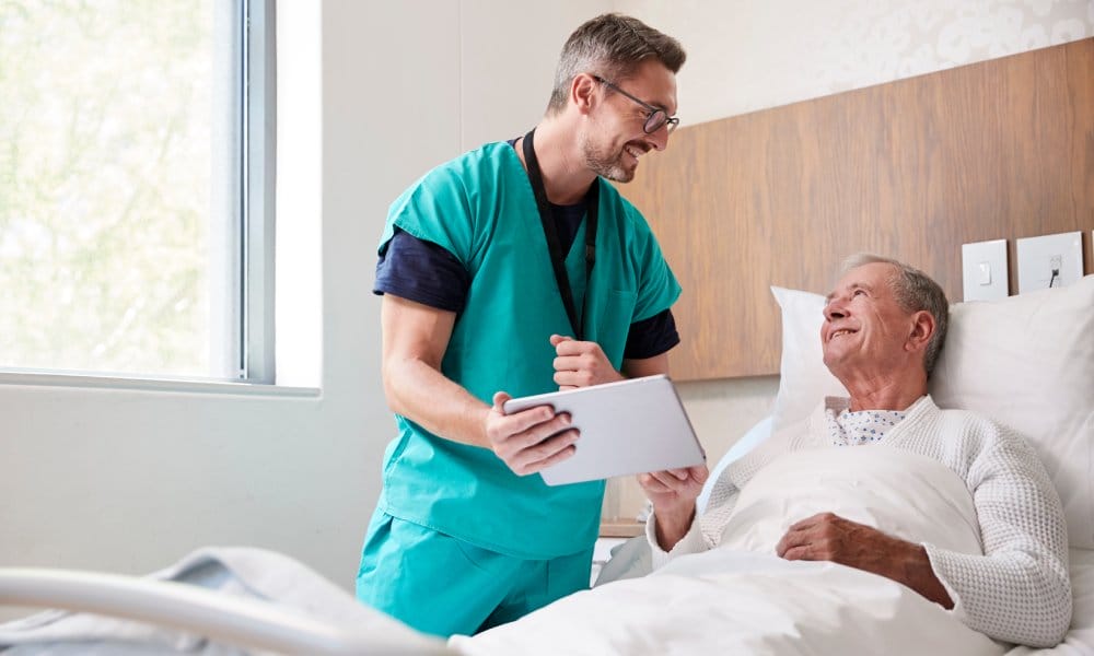 A medical professional checking up on his patient who is lying in bed. He is holding a tablet in front of his patient.