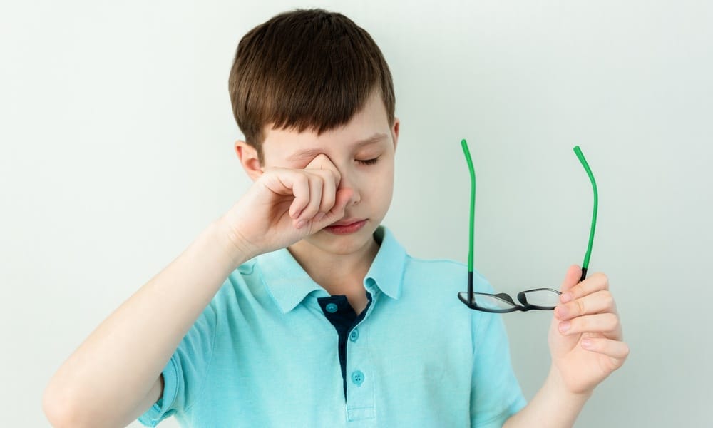 A young boy rubs his eye with one hand while holding his glasses in the other. He stands in front of a gray wall.