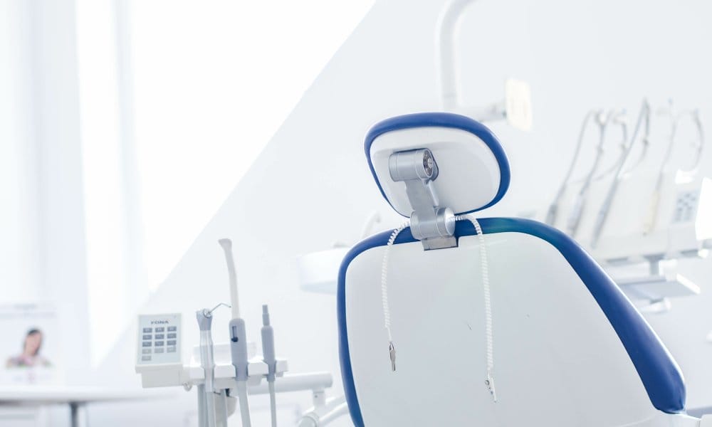 A dental practice treatment room with a white and blue patient chair facing away from the viewer next to tools and equipment.