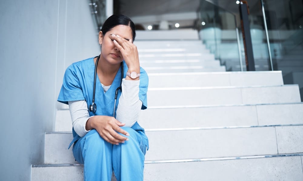 A woman wearing blue scrubs sits on white stairs. She has one hand sitting in her lap, and the other is rubbing her eyes.