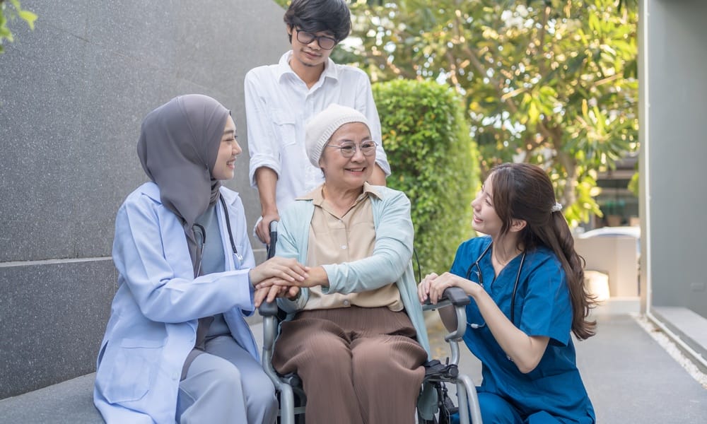 An older adult woman in a wheelchair wearing a headscarf. A supportive health-care team and family members surround her.