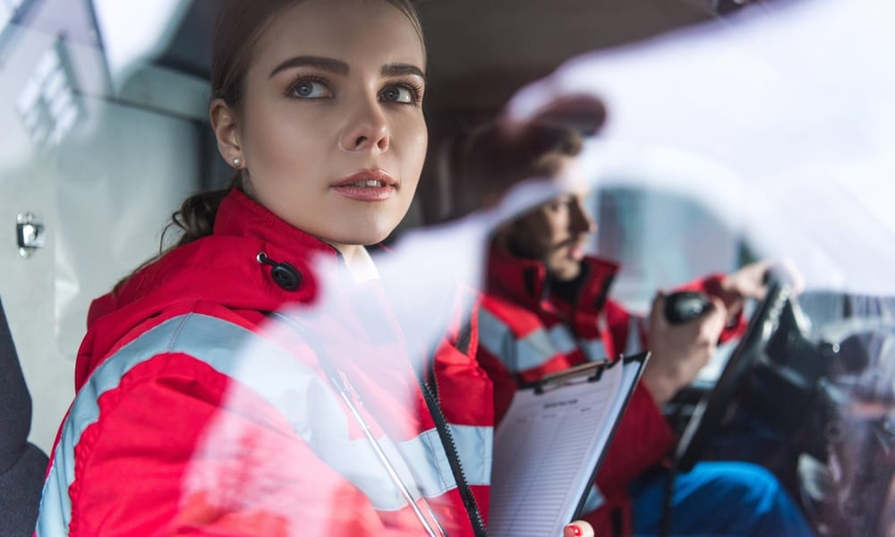 A female EMT sitting in an ambulance next to her driving coworker. She's holding a clipboard and looking out the window.