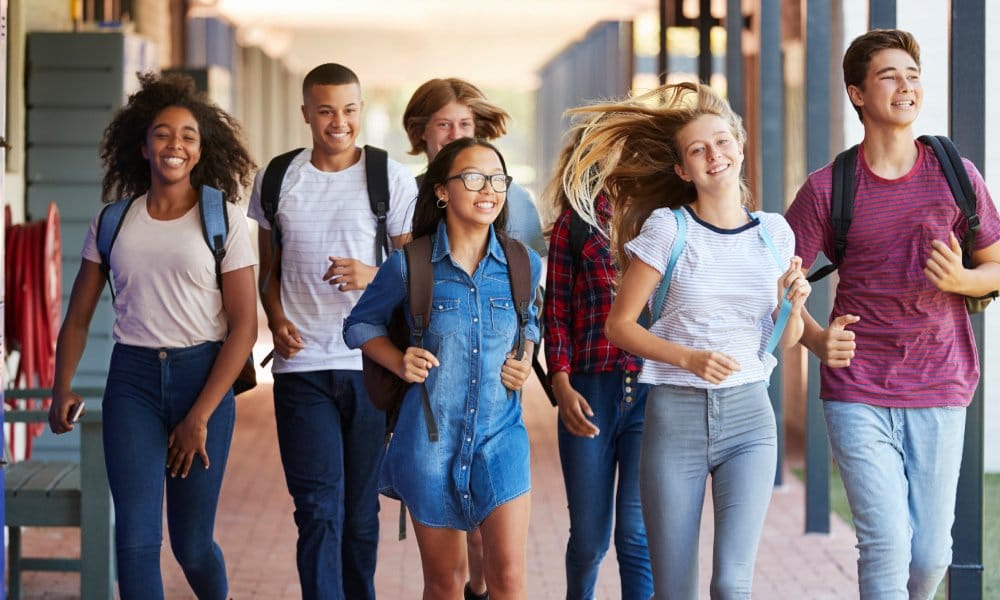 A group of smiling high school students wearing backpacks walk down the brick-floored hallway at school.