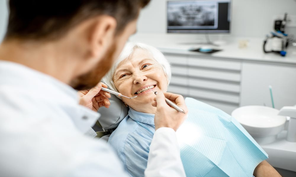 A senior woman sits in a dental chair and smiles at a dentist holding stainless steel tools near her mouth.