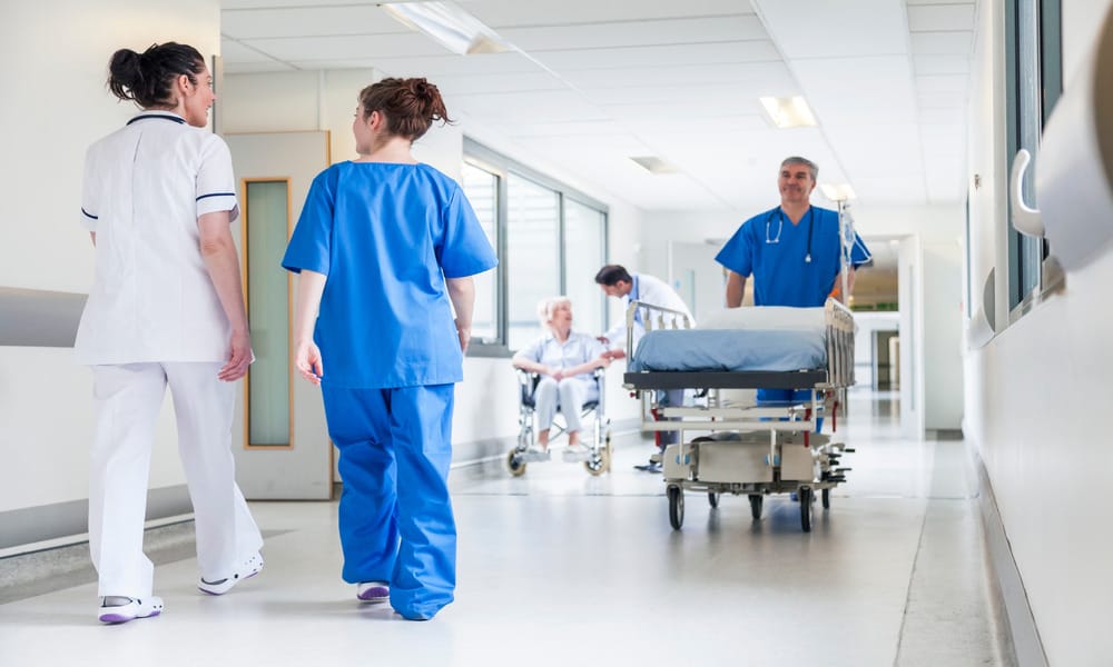 Multiple nurses in a hospital hallway walking, pushing a large rolling bed and talking to a senior patient in a wheelchair.