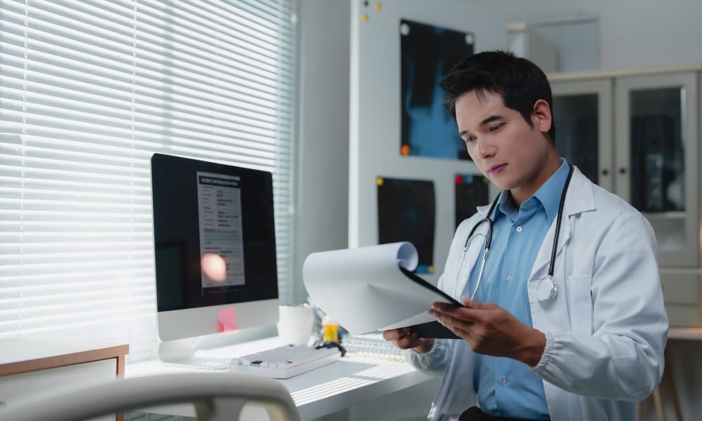 A male medical practitioner in a white lab coat is looking at a medical chart. There are X-rays in the background.
