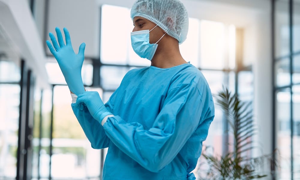 A healthcare professional pulls on a pair of blue gloves. They are also wearing a gown, mask, and hair covering.
