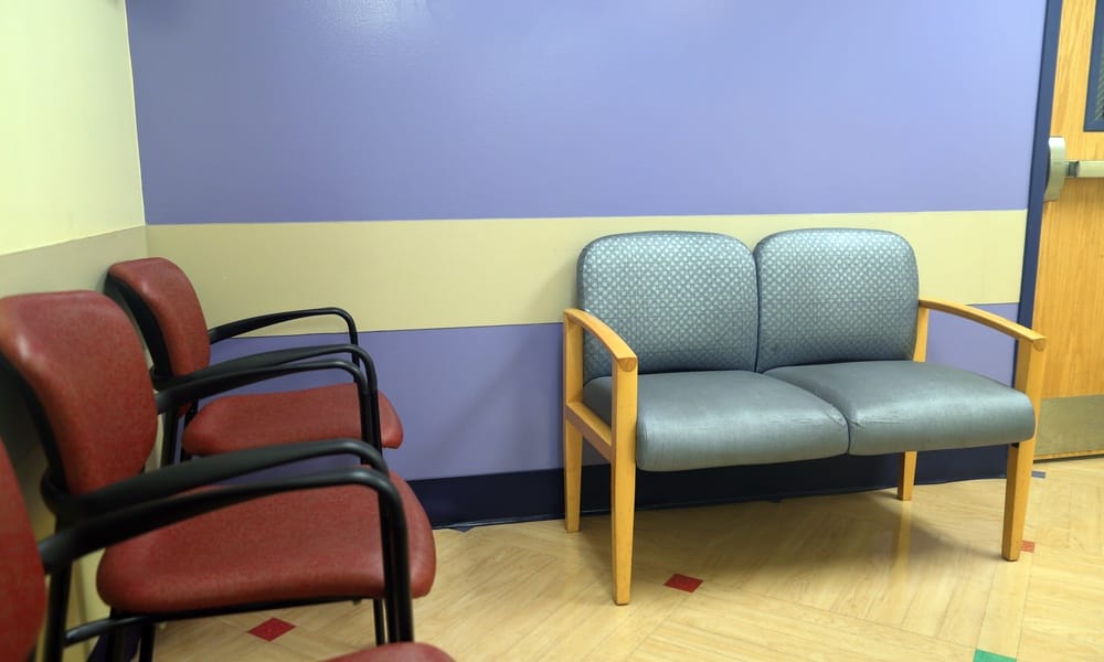 A beige clinic waiting room with a blue and white accent wall. Against a beige wall is a line of three red chairs.