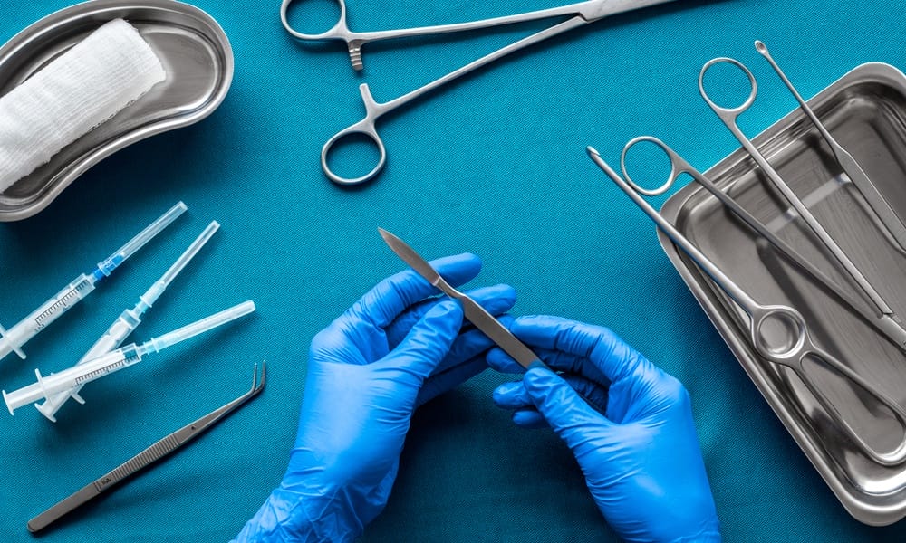 A surgeon at the tool tray wearing blue latex gloves and holding a surgical tool. There's other tools on the tray.