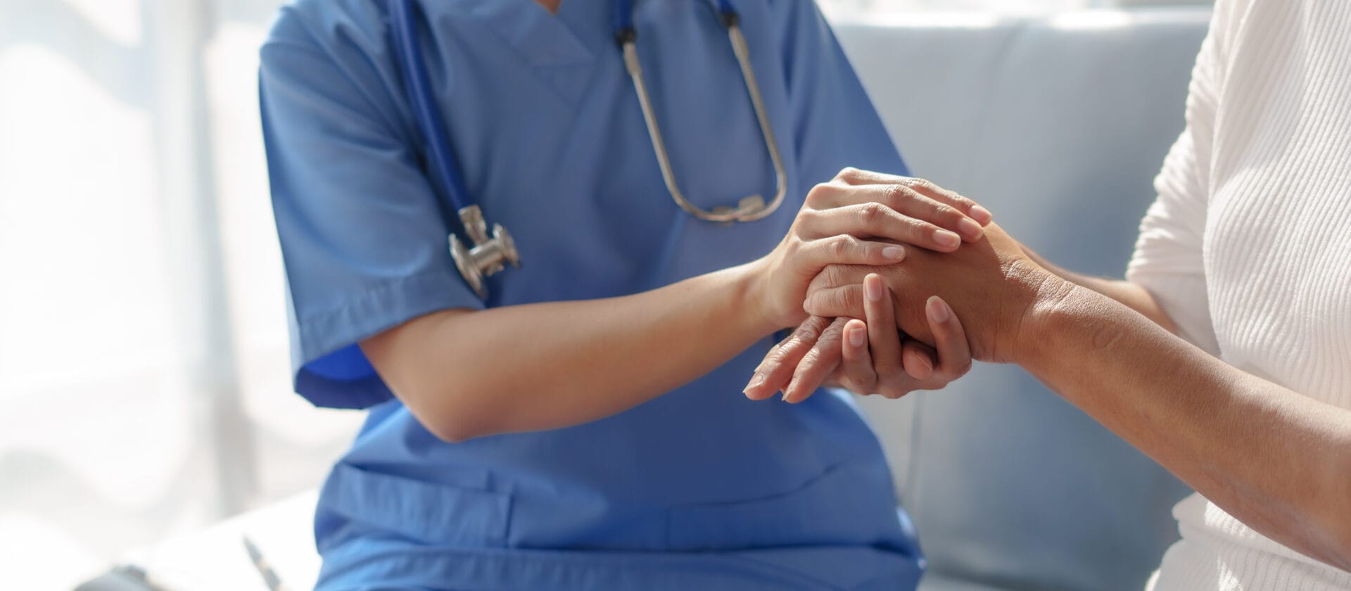 A healthcare professional in blue scrubs with a stethoscope around their neck is holding a patient’s hand.