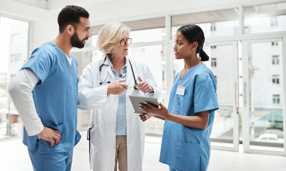 A senior doctor standing with a male and female nurse in blue scrubs in the hallway. The female nurse has a tablet.