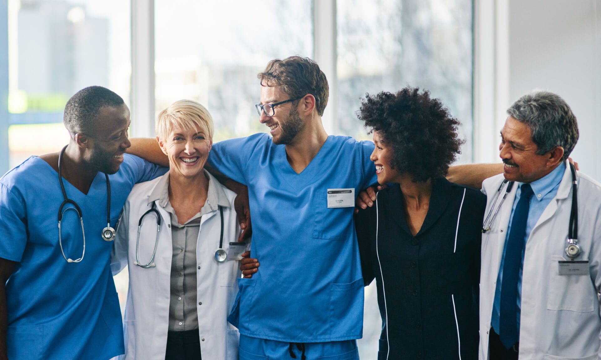 Five smiling healthcare professionals in scrubs and lab coats stand with their arms around each other as a team.