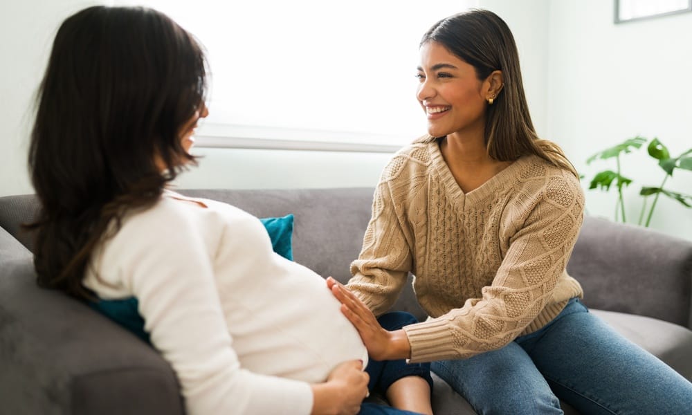 A professional midwife sitting with an expecting patient on a couch. The midwife is feeling the patient's belly and smiling.
