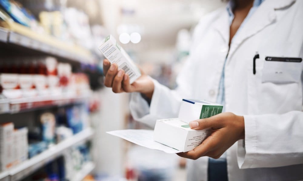 A pharmacist standing by a medicine storage cabinet holding a box of medication in their right hand and two in their left.
